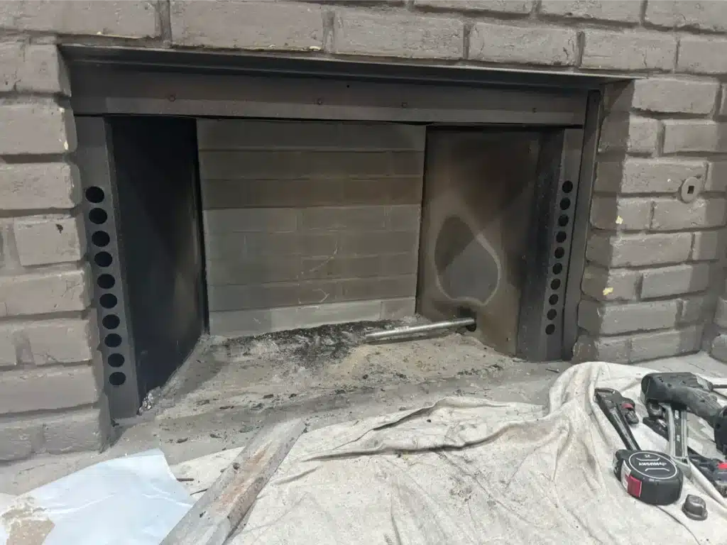 Open fireplace firebox with soot-stained brick and exposed metal air tubes during chimney damper repair.