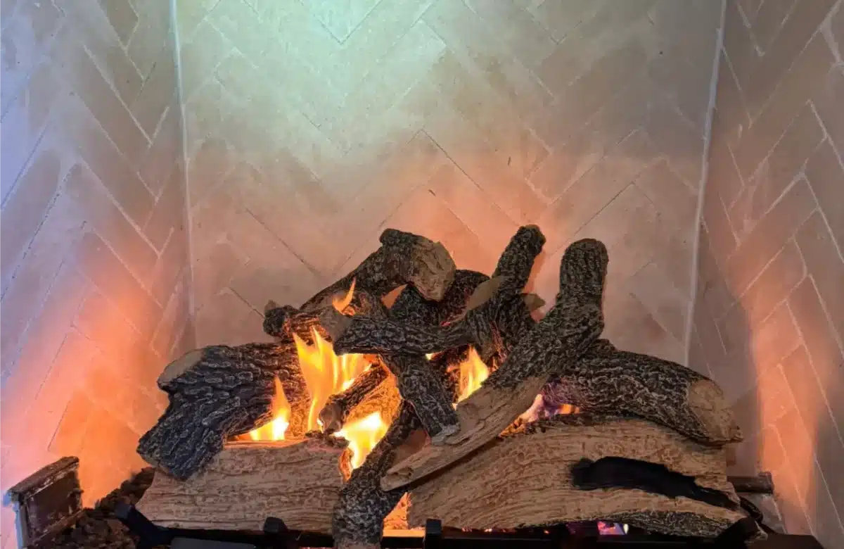 Burning logs resting on a black metal fireplace grate inside a herringbone brick firebox.