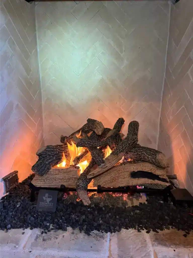 Burning logs resting on a black metal fireplace grate inside a herringbone brick firebox.