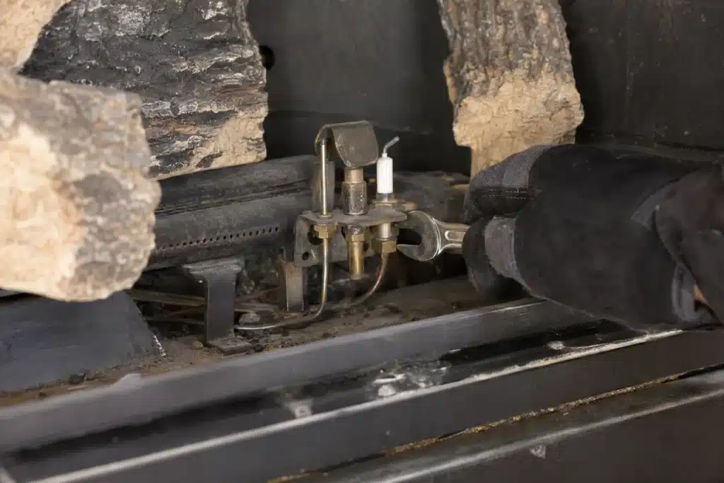 Close-up view inside an open gas fireplace firebox showing a pilot assembly - pilot hood, thermocouple rod, and igniter electrode - mounted on the burner rail, with a gloved hand holding a wrench nearby